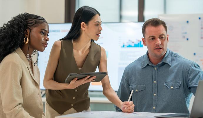 Three colleagues meet around a laptop in a casual office setting to discuss business strategy.