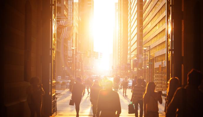 Pedestrians walk through a city street, surrounded by skyscrapers that frame the setting sun.