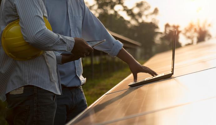 Two workers use a laptop and smartphone to inspect solar panels at sunset