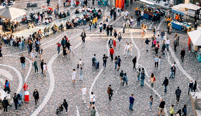 An overhead view of a public square, patterned with stone tiles, shows people walking, gathering, and sitting amid a fountain and market stalls.