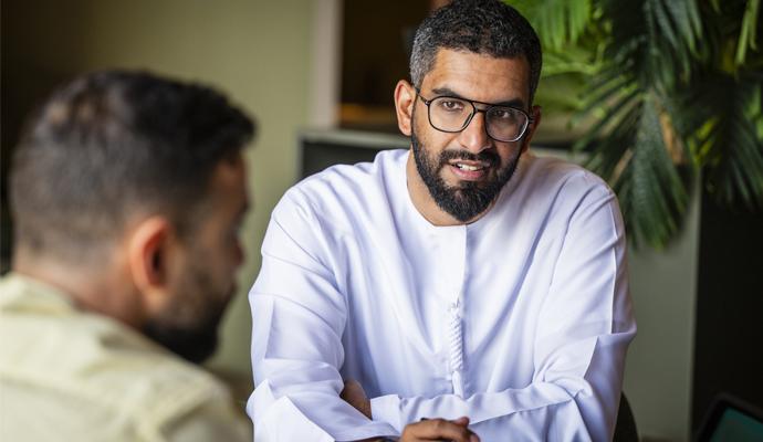 A businessman wearing a white shirt and glasses sits at a table, conversing with a colleague.