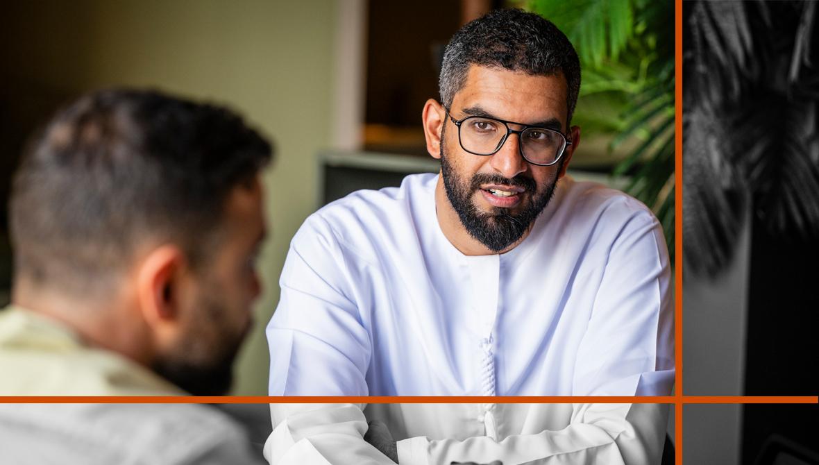 A businessman wearing a white shirt and glasses sits at a table, conversing with a colleague.