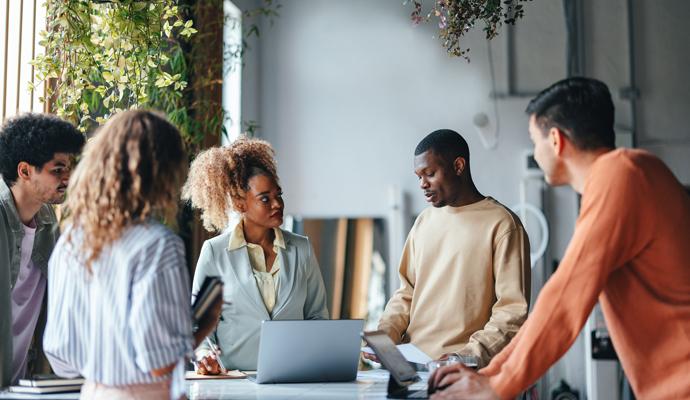 A diverse group of colleagues stands and meets around a table in an informal office environment.