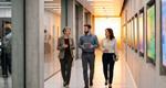 Three professionals converse while walking along a hallway in a modern office building with an industrial design. On the left of the image, the hall is bordered by glass-paneled meeting rooms; on the right, there's a wall with video monitors.