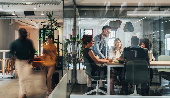 Five coworkers meet around a table inside a glass-walled conference room as colleagues walk by in the open office outside.