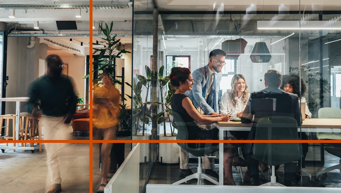 Five coworkers meet around a table inside a glass-walled conference room as colleagues walk by in the open office outside.