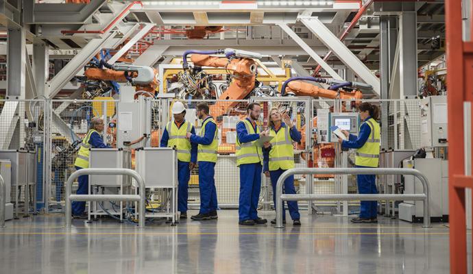 A team of workers in safety vests stands on a manufacturing floor in front of robotic machinery.