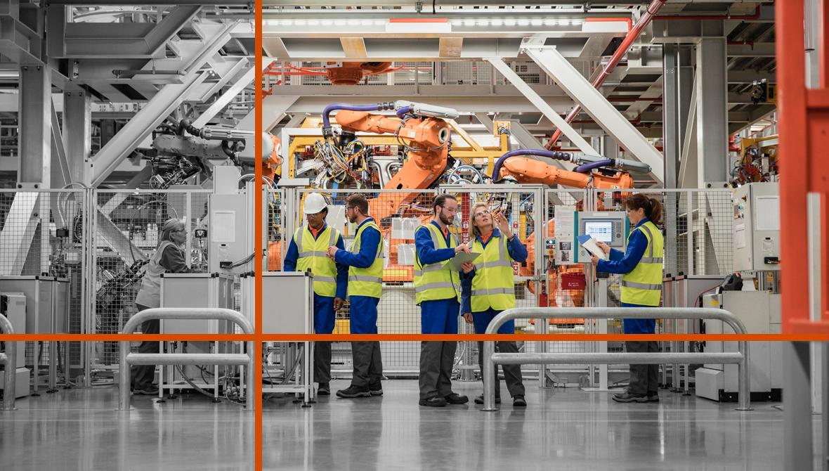 A team of workers in safety vests stands on a manufacturing floor in front of robotic machinery.