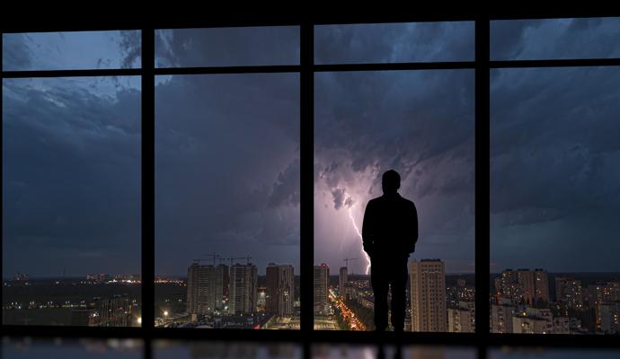 A businessman facing a panoramic window at nighttime with a bolt of lightning in the background