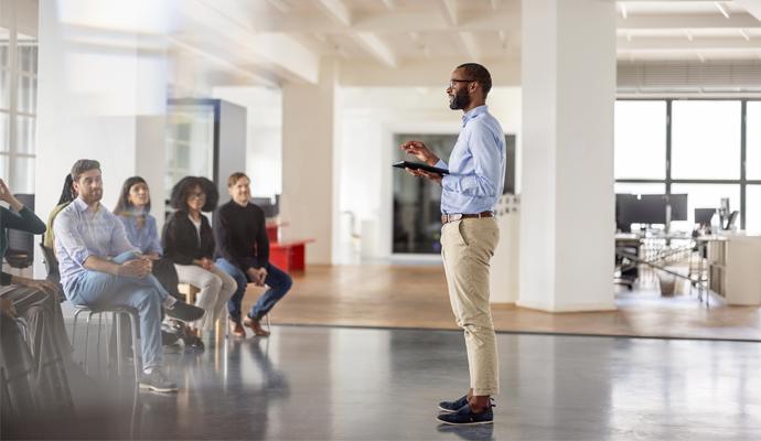 : A businessman stands and addresses his seated colleagues with a digital tablet in hand.