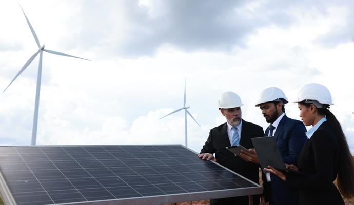 Three inspectors in suits and hard hats look at a solar panel with wind turbines in the background