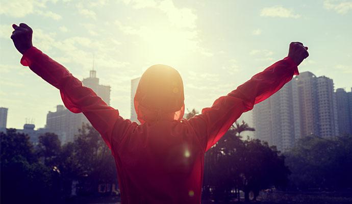 Woman overlooking a city at sunrise raises her arms overhead triumphantly