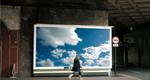 A woman turns to look at a large billboard image of white clouds in a blue sky while walking along a sidewalk.