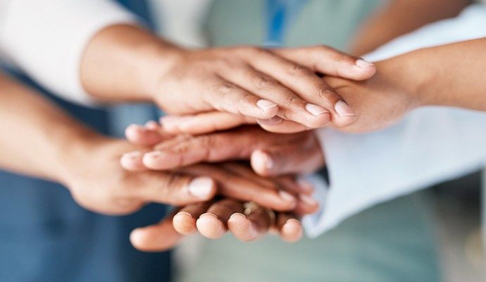A close-up photo of stacked hands in the middle of a group of businesspeople suggests teamwork and collaboration