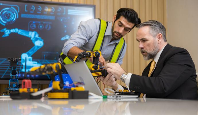 Two engineers program a robotic arm while an image of the arm appears on a monitor behind them