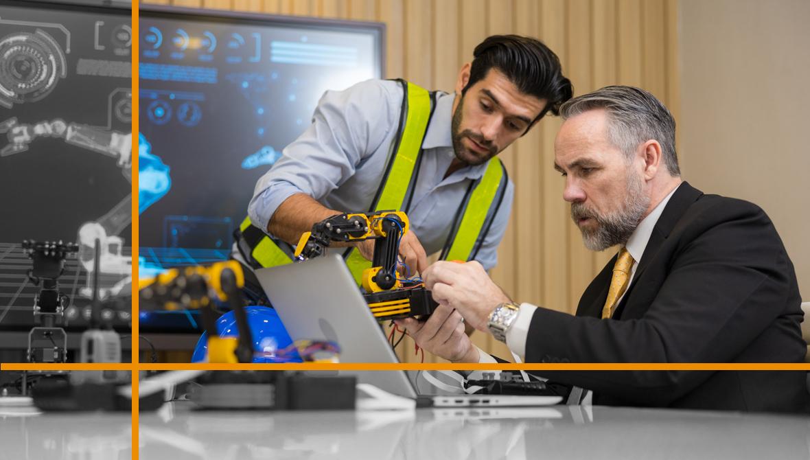Two engineers program a robotic arm while an image of the arm appears on a monitor behind them