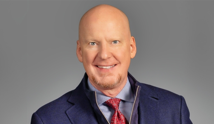 A photo of John Riggs wearing a blue suit jacket over a blue checked shirt and red tie, against a gray background