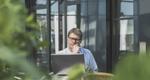 Photo of a man sitting outside with a laptop, with shrubbery in the foreground 