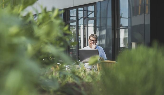 Photo of a man sitting outside with a laptop, with shrubbery in the foreground  Photo of a man sitting outside with a laptop, with shrubbery in the foreground