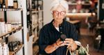 A woman with white hair and red-framed glasses holds a reusable bag while examining a packaged product in a grocery store.