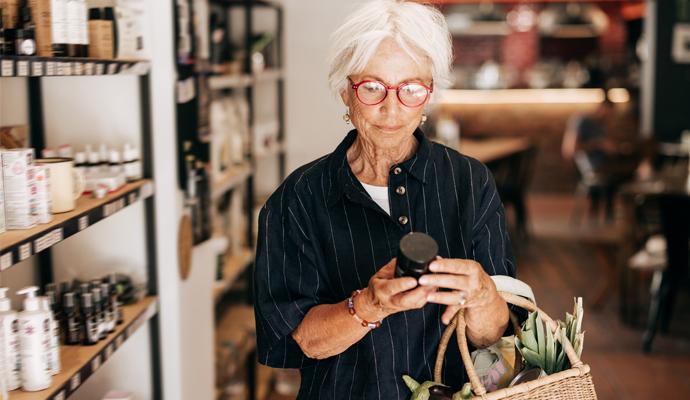 A woman with white hair and red-framed glasses holds a reusable bag while examining a packaged product in a grocery store.