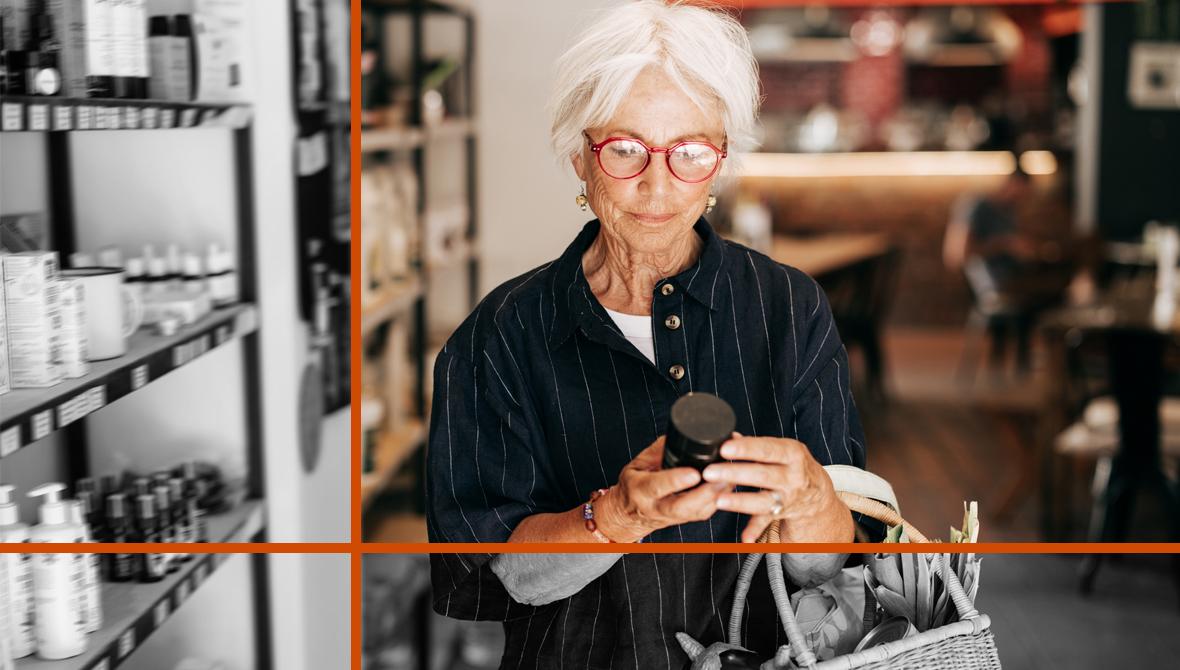 A woman with white hair and red-framed glasses holds a reusable bag while examining a packaged product in a grocery store.