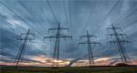 Rows of high-voltage power lines stretch across a field under a cloudy sky at sunset.