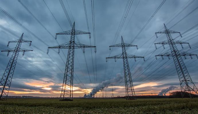 Rows of high-voltage power lines stretch across a field under a cloudy sky at sunset.