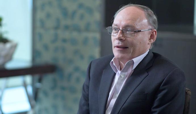 Pollitt, wearing glasses, a white shirt, and a gray suit jacket, seated in an office conference room