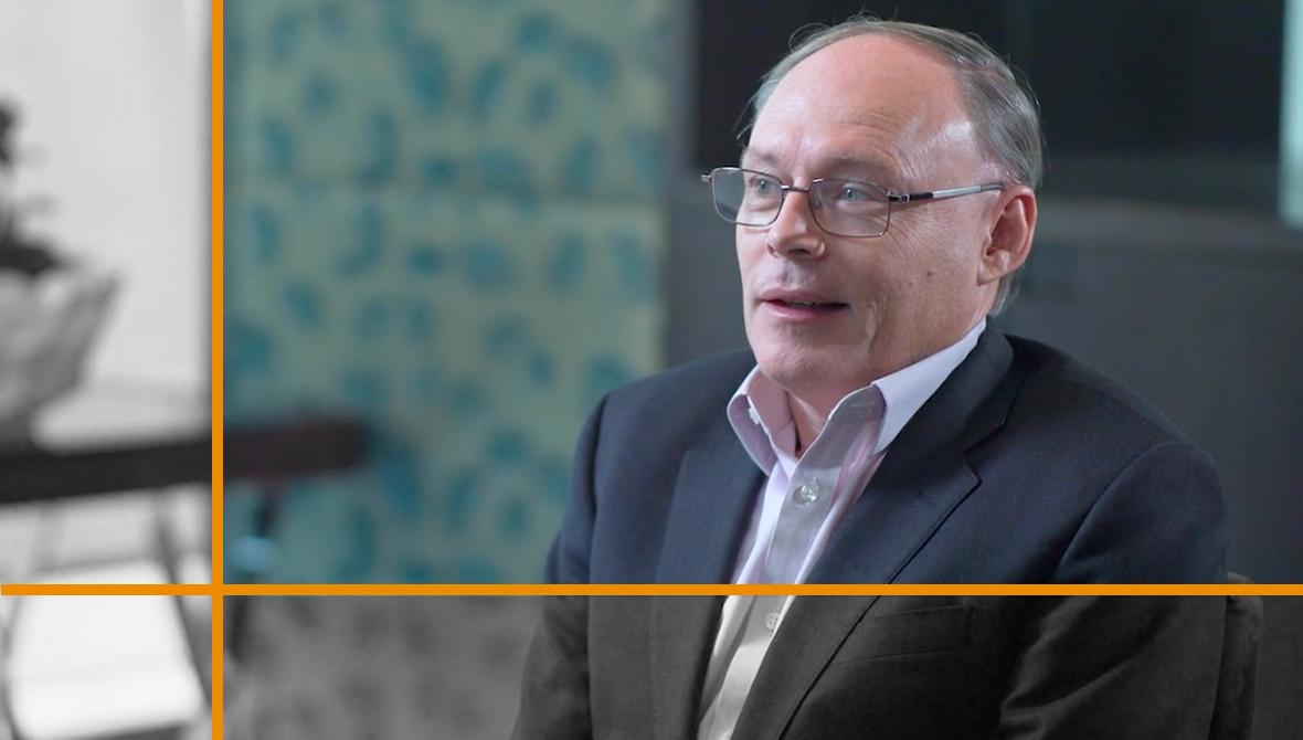 Pollitt, wearing glasses, a white shirt, and a gray suit jacket, seated in an office conference room