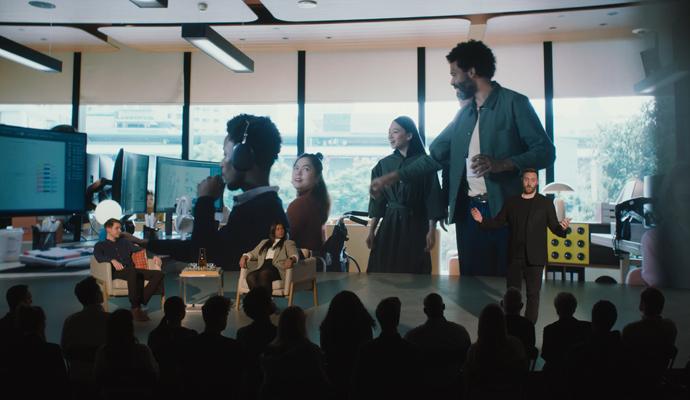 Stage presentation with seated panelists and a presenter, as an audience watches and a large screen shows coworkers in an office.