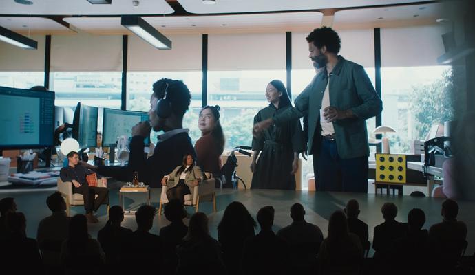 Stage presentation with seated panelists and a presenter, as an audience watches and a large screen shows coworkers in an office.