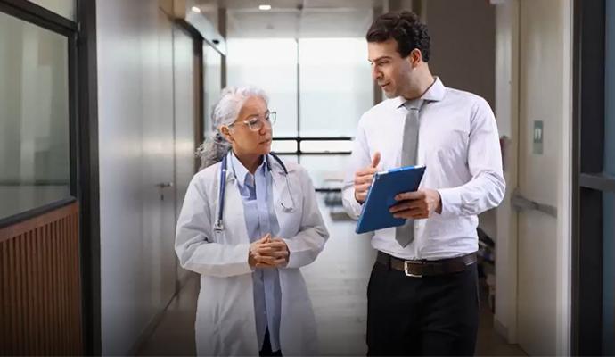 A doctor walks through a hospital corridor while speaking with a colleague holding a clipboard.
