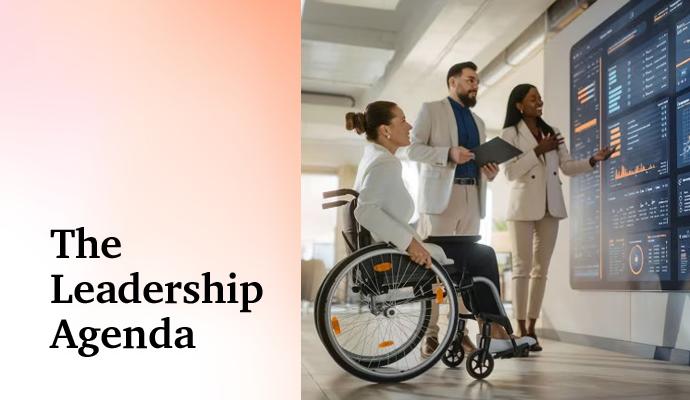 Team of colleagues standing and seated in a wheelchair, discussing charts displayed on a wall monitor.
