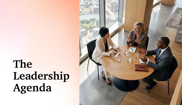 Three professionals engage in conversation at a round table in a modern office with a city view behind them.